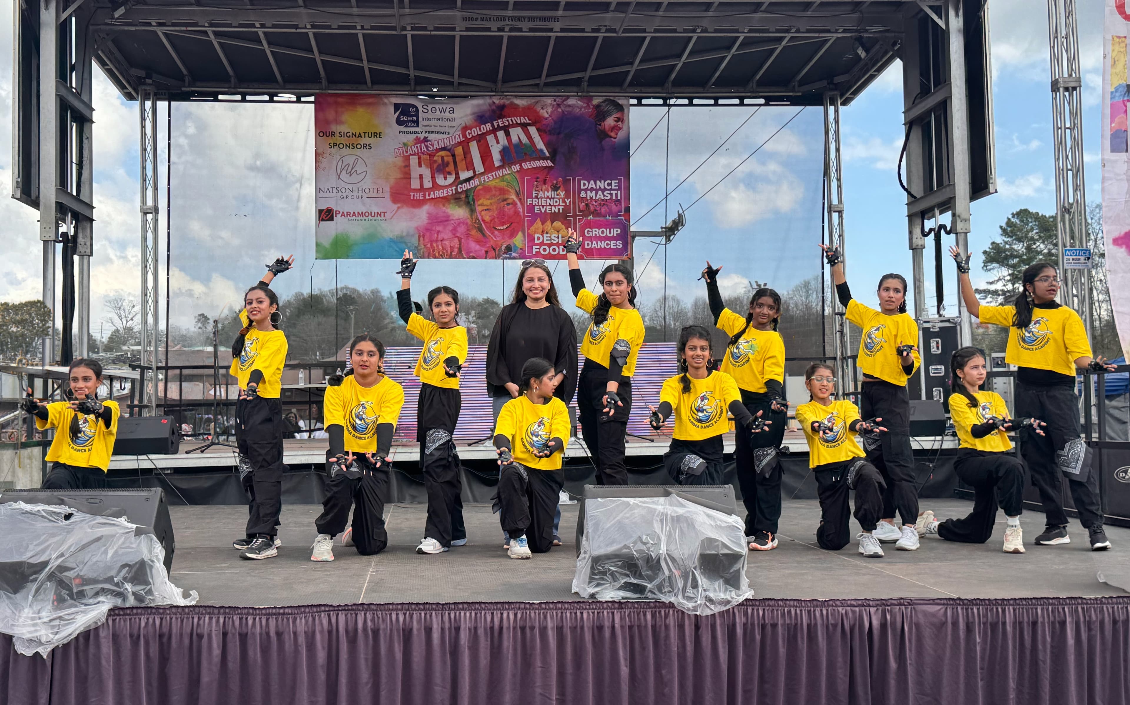 Garima Dance Academy group with the instructor in matching yellow tops on a Holi festival outdoor stage, arms raised
