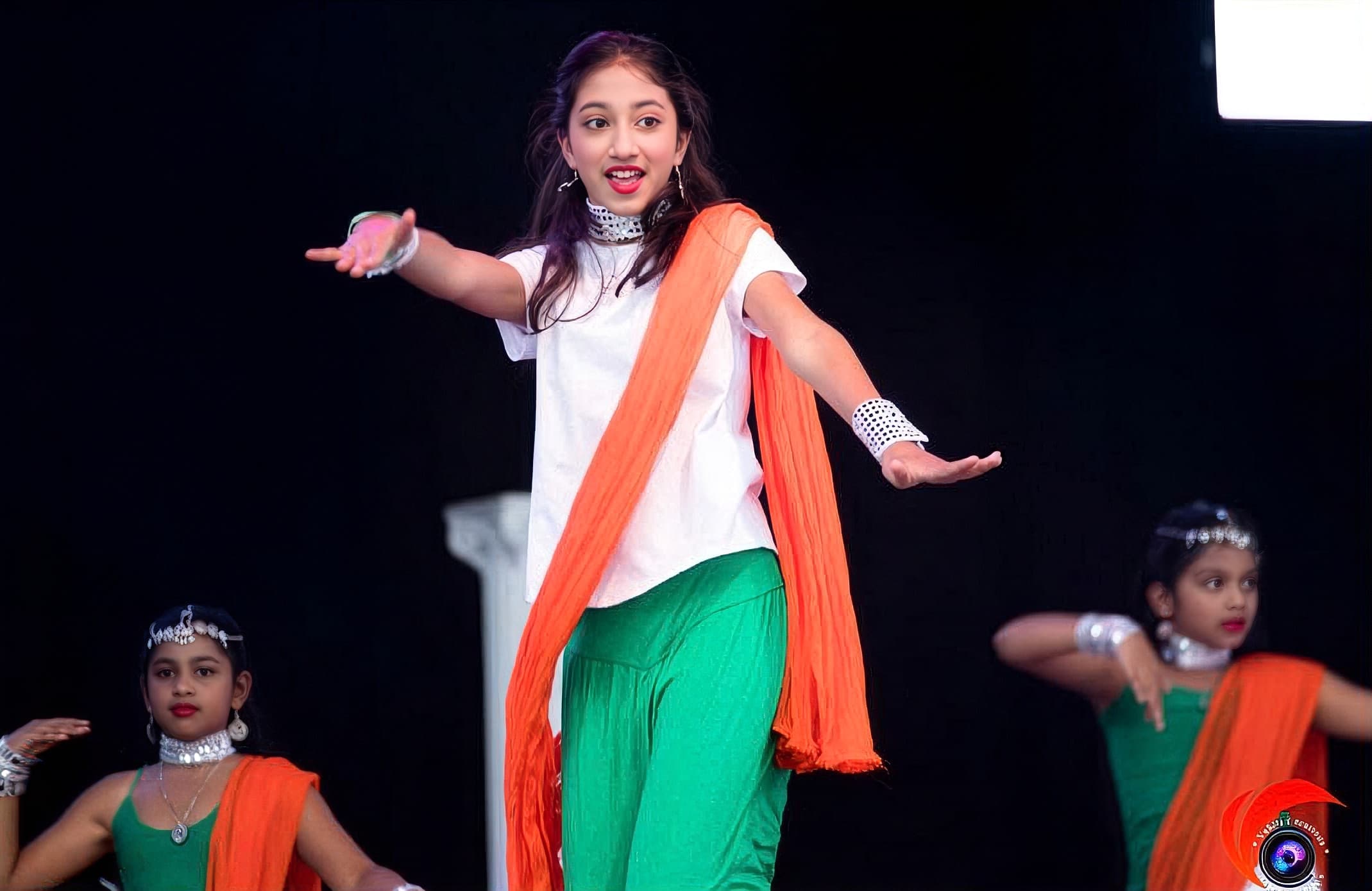 Solo dancer in white, orange, and green flag-color attire mid-performance with arms outstretched on a dark stage