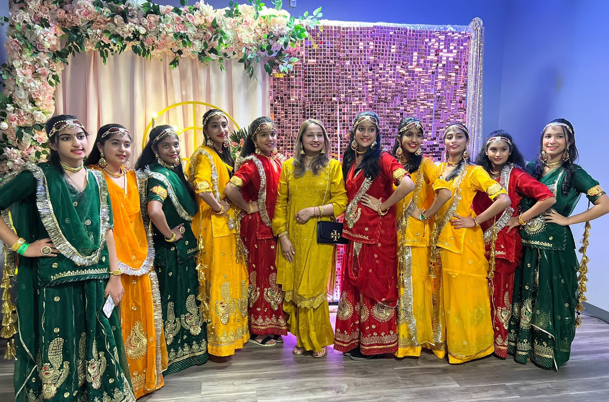 Teen students with the instructor in red, yellow, and green traditional attire, posed in front of a floral and sequin backdrop